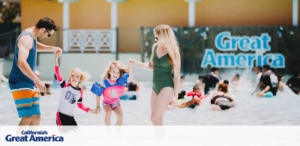 Image shows a joyful moment at California's Great America. Two excited children, wearing swimwear and rash guards, playfully hold hands with an adult male in swim trunks. Another adult female in a green swimsuit walks by the shallow water area where several guests enjoy the water. The park's name is visible in the background, evoking a lively, family-friendly atmosphere.