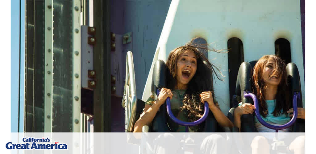 Image shows two people on a roller coaster ride at 'California's Great America' amusement park. Their expressions are of excitement and thrill, with their mouths open as they hold on to the safety harness. Bright sunlight enhances this dynamic, joyful moment.