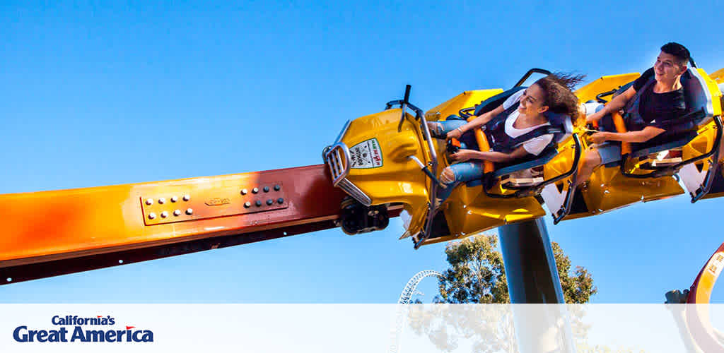 Image shows a thrilling ride at California's Great America theme park with two excited riders in a yellow cart that simulates flying, against a clear blue sky. The ride's arm extends from the left with the Great America logo visible.