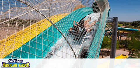 A thrilling scene at Six Flags Hurricane Harbor where visitors are enjoying a water slide. The slide is enclosed, with blue and yellow colors, twisting downwards. People are caught mid-descent in a translucent section, their arms raised in excitement. The background reveals a sunny day with clear skies and additional water attractions.