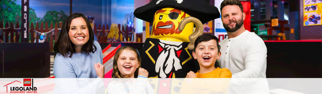 This image features a joyful family of four at a LEGOLAND Discovery Center, smiling widely and posing for a photo. On the left, a woman with long dark hair and a light blue top stands beside a young girl with shoulder-length hair who is wearing a bright red shirt. To their right, a young boy in a yellow shirt is laughing, with a man sporting a short beard and white long-sleeve shirt standing behind him. Next to the family stands a life-size model of a LEGO pirate, complete with a black hat with a skull and crossbones emblem. They are all posed before a colorful background that resembles a LEGO-themed play area filled with vibrant details. The LEGOLAND Discovery Center logo is visible in the bottom left corner.

FunEx.com is dedicated to bringing families unforgettable experiences with the added bonus of savings—find the lowest prices on tickets to enchanting destinations like LEGOLAND Discovery Centers!