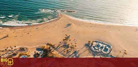 This image features a scenic aerial view of a serene beach during what appears to be the late afternoon. The golden sand creates a wide border along the curve of the coastline, leading into the gentle, blue waves of the sea. On the beach, there are several umbrellas casting long shadows, suggesting the presence of a leisurely atmosphere. A small jetty extends into the water, and to the right, there is a circular structure with a pool surrounded by lounging areas. The perspective is from above, providing a grand vista of the beach and its tranquil setting. The clear weather and sunny conditions contribute to the inviting feel of the scene.

At FunEx.com, we're committed to ensuring you get the lowest prices and hefty savings on tickets to your favorite destinations, just like the picturesque beach featured in this image.