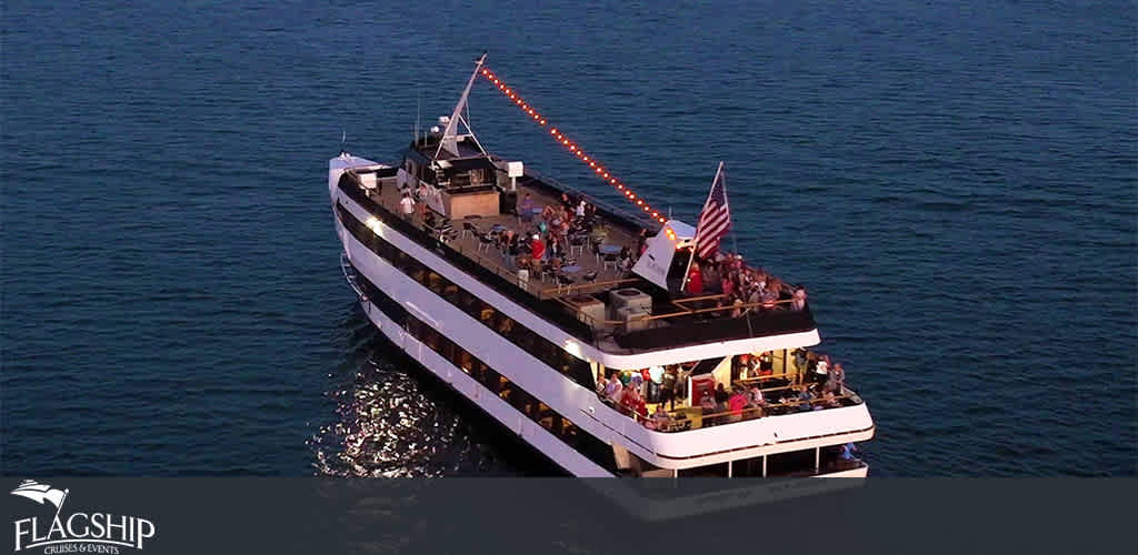 This image displays a large multi-deck cruise ship belonging to Flagship Cruises & Events, floating peacefully on a calm body of water during what appears to be dusk, given the dim light. We can observe an American flag prominently displayed at the rear of the upper deck. The vessel features string lights that add to the ambiance and several people can be seen enjoying their time on both the upper open deck and the illuminated decks below. A silhouette of the coastline is faintly visible in the background, suggesting the ship is not far from shore.

Be sure to check out FunEx.com for exclusive discounts and the lowest prices on tickets for your next aquatic adventure.