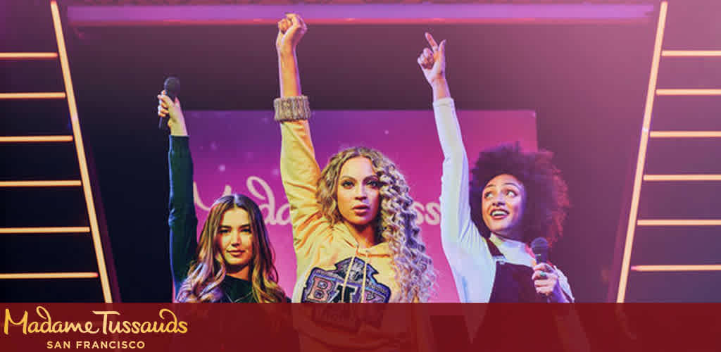 This image features a vibrant display from Madame Tussauds San Francisco, showcasing three lifelike wax figures that resemble iconic music artists. From left to right, there's a figure with long, light brown hair and a black leather jacket, another in the center with long curly blonde hair and a blue and gold varsity jacket, and the figure on the right sporting a voluminous curly afro and a white blouse. Each figure is striking a dynamic pose, with the left figure holding a microphone in her right hand, the middle one raising her right hand high with a peace sign, and the right figure holding a microphone in her left hand and her right hand outstretched upward. The energy and excitement of a live performance are captured in their expressions and gestures. The setting is illuminated with magenta and yellow stage lights, contributing to the lively atmosphere. In the foreground, the famous Madame Tussauds logo is prominently displayed, indicating the curated exhibition these figures are a part of.

At FunEx.com, not only do we offer you a glimpse into such thrilling experiences, but we also guarantee savings with the lowest prices on tickets to make your visit even more enjoyable.