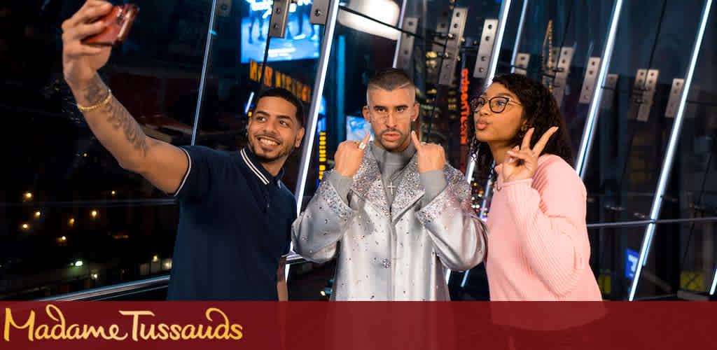 Image Description: This image captures three individuals enjoying a moment together in front of a backdrop with the "Madame Tussauds" logo. On the left side, a man with short hair, a tattoo on his left arm, and wearing a dark polo shirt, smiles widely as he takes a selfie. In the center, an individual wearing a shiny, silver, bedazzled jacket with a serious expression is making a fist with their hands near their chin, as though striking a boxing pose. On the right, a woman with curly hair wearing glasses and a pink sweater poses with a peace sign. The group appears to be standing in front of a mirrored surface that reflects bright, linear lights, likely inside the famous wax museum at night. Explore the wonders of Madame Tussauds and share a memorable experience with friends—don't forget to visit FunEx.com for the lowest prices on tickets and additional savings!