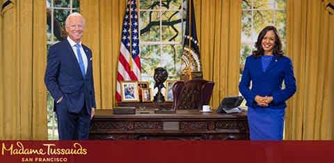 This image shows two lifelike wax figures displayed in an office setting reminiscent of the Oval Office. To the left stands a male figure posed with one hand in his pocket and a slight smile. He is dressed in a dark suit with a blue tie. To the right, a female figure is seen with a wide smile, standing confidently, hands crossed in front of her, and dressed in a business-like blue dress. Behind them, an American flag and a desk with several items on it, including a phone, papers, and a bronze eagle sculpture, are visible. A plaque at the bottom of the image indicates that this is an exhibit at Madame Tussauds San Francisco. Incorporating fun experiences into your life doesn't have to break the bank—visit FunEx.com for the lowest prices on tickets and amazing savings on attractions.