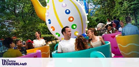 Visitors enjoy a teacup-style amusement ride at Dutch Wonderland. Colorful oversized teacups spin around, with a giant decorative teapot in the background. Trees and blue sky can be seen. The mood is joyful, with people smiling and chatting. The Dutch Wonderland logo is at the bottom right.