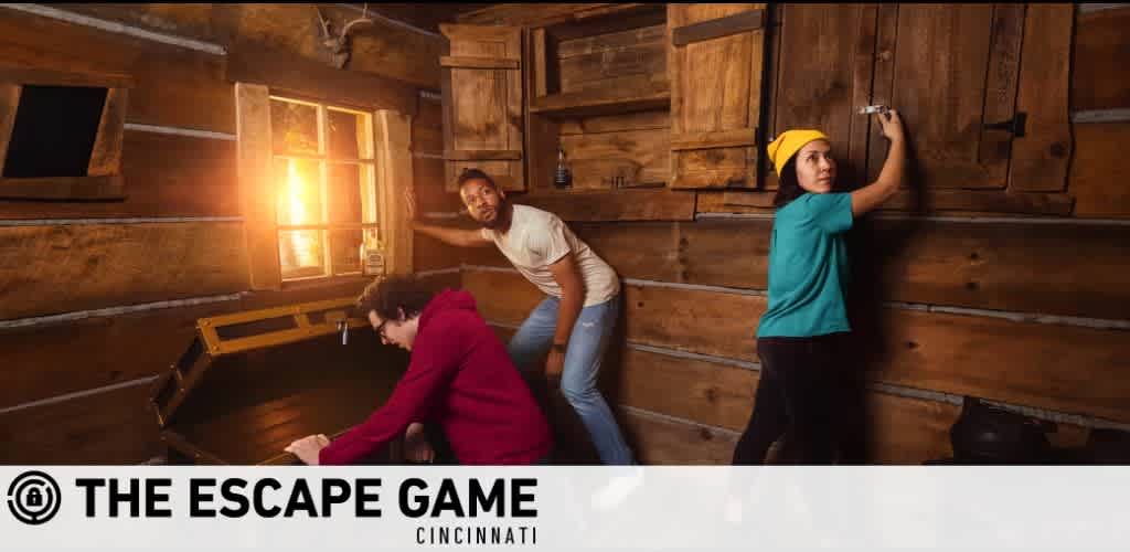 Image shows three people engaged in a puzzle-solving activity at The Escape Game Cincinnati. A person in red is bending over an open chest, a person in white looks on with interest, and a person in teal is standing and reaching towards a latch on a wooden wall. The room is warmly lit and has a rustic cabin feel.