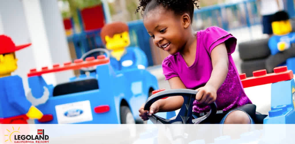 A joyful young girl with her hair styled in curls is wearing a fuchsia dress as she pretends to drive a blue LEGO car with a delighted expression at the LEGOLAND California Resort. In the background, oversized LEGO figures add to the playful setting. The LEGOLAND logo graces the bottom of the image.