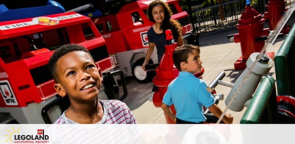 Image of joyful children at LEGOLAND California Resort fire station attraction. A boy in the foreground looks upward with a smile, while another child operates a firetruck ride apparatus. A girl in the background stands beside a red, LEGO-style firetruck, also smiling. Bright sunlight and vibrant colors convey a fun, energetic atmosphere.