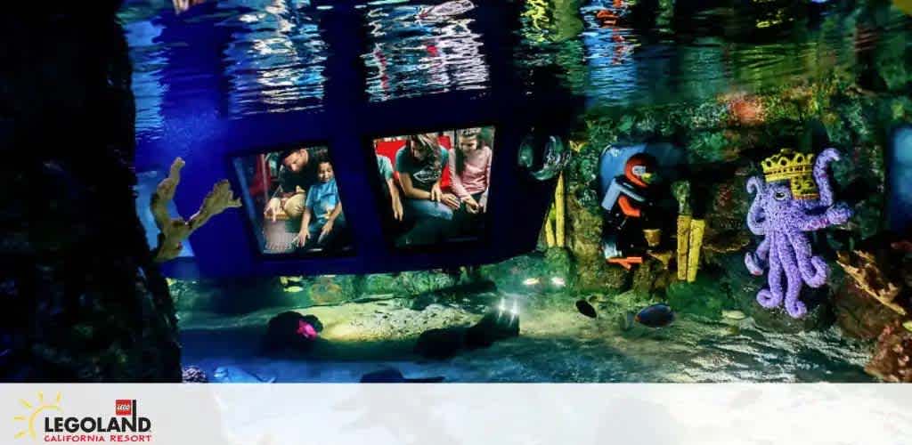 Image shows an underwater adventure scene at LEGOLAND California Resort. Visitors are inside a submerged vehicle with large viewing windows, observing colorful LEGO figures such as a diver and a sea creature, amidst a vibrant artificial coral reef with scattered LEGO bricks.