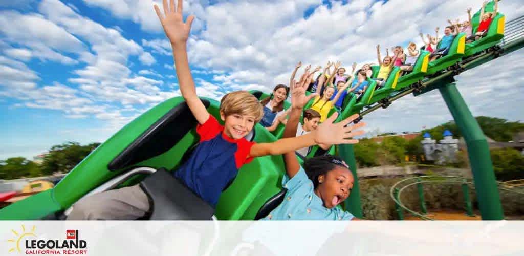 Visitors enjoy a thrilling roller coaster ride at LEGOLAND California Resort. They are smiling and raising their hands in excitement as their green coaster cart races down the track. The backdrop features a clear blue sky with wisps of white clouds.