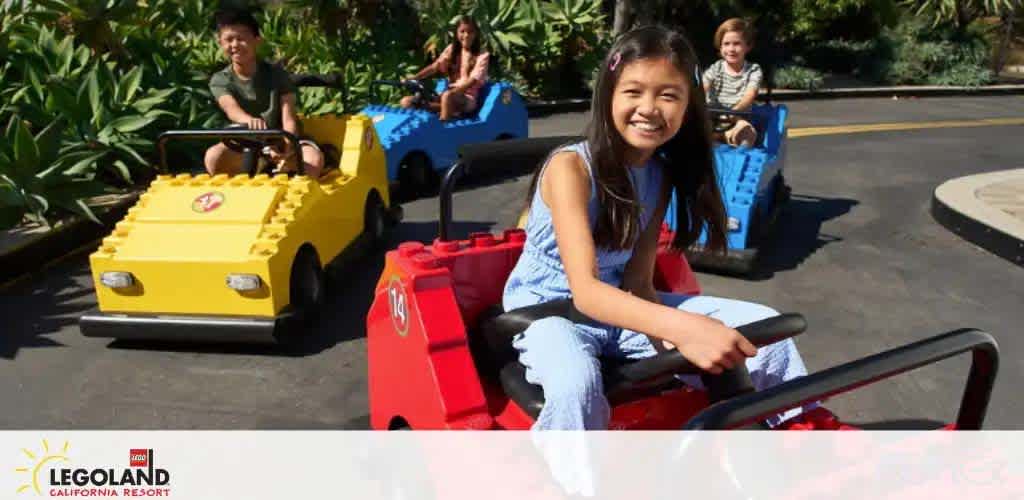 At LEGOLAND California Resort, children enjoy a sunny day driving colorful, life-sized Lego cars. A joyful girl in a blue dress pilots a red car in the foreground, while others steer yellow and blue cars behind her, amid lush greenery.