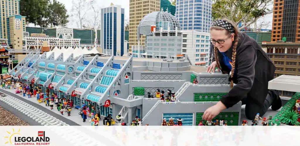 A person is interacting with a detailed LEGO model of a cityscape at LEGOLAND California Resort. The display includes miniature buildings, trains, and LEGO figures. The individual is focused on adjusting a section of the model. Trees and vibrant signs complement the busy layout.