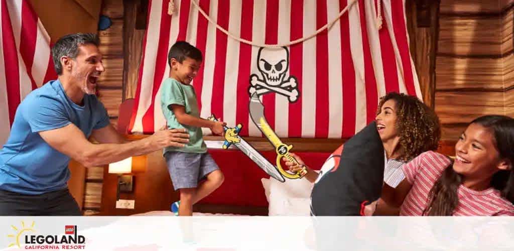 In a pirate-themed room at LEGOLAND California Resort, a man and three kids engage playfully. The man pretends to sword fight with a boy, while two girls laugh, clutching a pillow with a skull and crossbones. The backdrop features a red and white striped curtain and a wooden cabin interior.