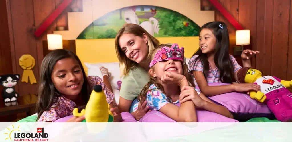 Image depicts a joyful moment at LEGOLAND California Resort with four individuals, possibly a family, interacting playfully. An adult woman and three young girls are seen on a colorfully made bed, surrounded by themed decorations and plush toys representing LEGO characters. The atmosphere suggests a fun, child-friendly environment.