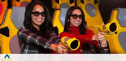 Two women with 3D glasses play an arcade shooting game, smiling.