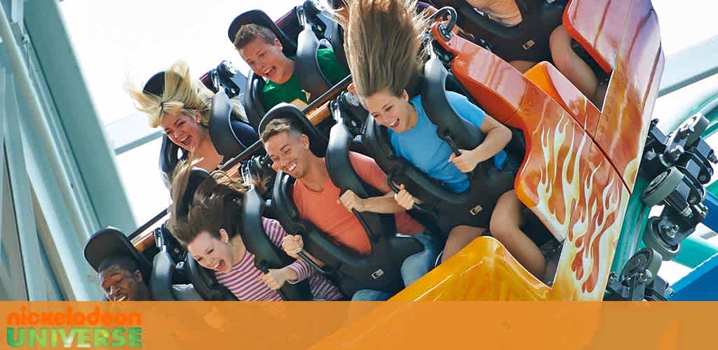 A group of excited riders experience the thrill of a roller coaster at Nickelodeon Universe. Their expressions range from wide smiles to exhilarated screams as they descend a track. The coaster car's flame design adds to the sense of speed. Bright sunlight illuminates the scene, capturing the vibrant atmosphere of the amusement park.