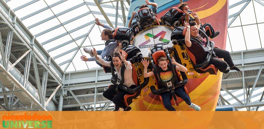 Thrilled riders are shown on an amusement park attraction at Nickelodeon Universe. They are secured in their seats with safety harnesses, hands raised in excitement, against a background of a colorful ride structure and a glass ceiling letting in natural light.