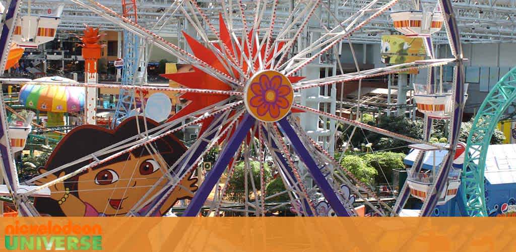 Image of an indoor amusement park featuring a vibrant Ferris wheel adorned with a colorful flower design at its center. Below, a large character mural adds to the playful atmosphere. The space is filled with bright structures and festive decorations, creating an inviting and fun-filled environment. A banner showcasing 'Nickelodeon Universe' indicates the themed nature of the park.