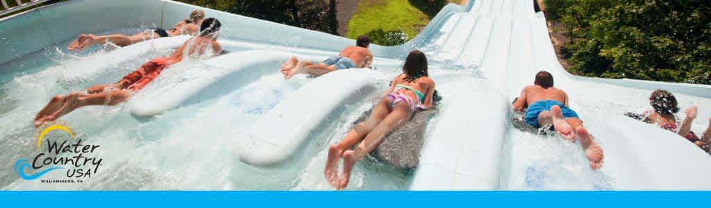 A group of people are enjoying a sunny day at a water park, sliding down a wide, multi-lane water slide. The slide is white with water flowing down it. There's foliage on the sides, and a logo in the lower left corner reads 'Water Country USA, Williamsburg, VA'.