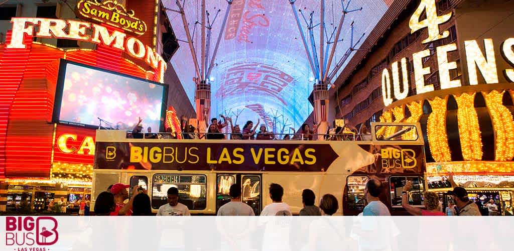 The image features the vibrant Fremont Street Experience in Las Vegas with a Big Bus tour vehicle in the foreground. Neon signs illuminate the area, highlighting attractions like the Four Queens and Fremont casinos. People are visible enjoying the bustling environment. The sky above is covered by a canopy screen, displaying colorful graphics. The Big Bus has branding on the side and a group of passengers on the open top deck.