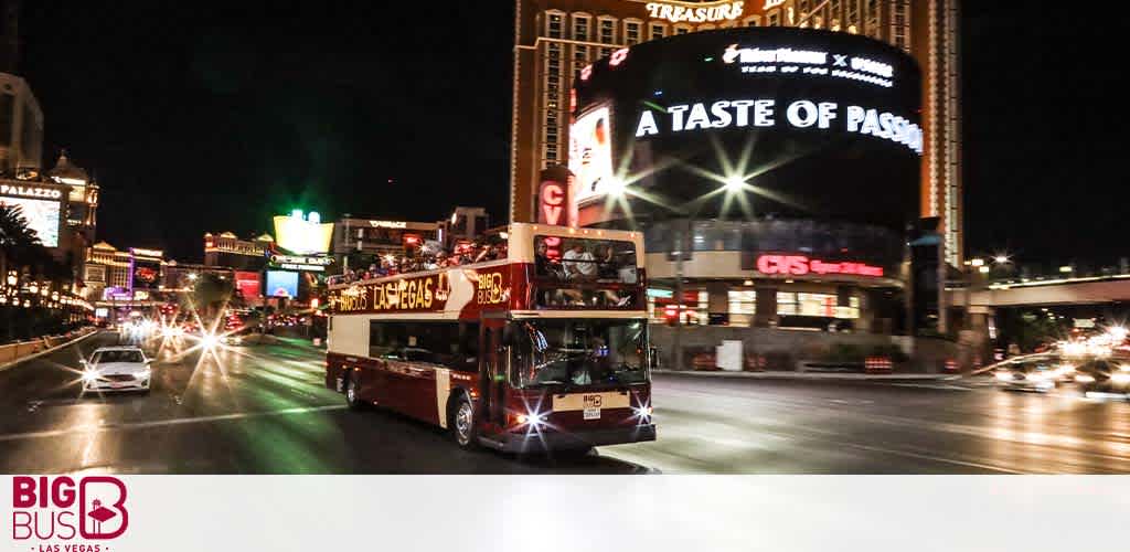 A red, double-decker tour bus labeled 'Big Bus Las Vegas' travels at night along a bustling street. Bright city lights and illuminated signs from nearby casinos and hotels, including the Treasure Island and Palazzo, create a vibrant atmosphere typical of Las Vegas. The scene captures the dynamic energy of the city.