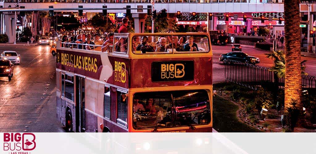 Image of a red double-decker Big Bus Las Vegas touring at dusk, filled with passengers on the upper open deck. City lights begin to illuminate the evening sky as the bus travels down a bustling street lined with palm trees and neon-lit buildings.