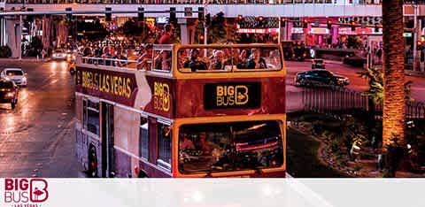 An open-top red double-decker Big Bus tours at twilight with passengers on the upper deck, cruising down a bustling street lined with palm trees and illuminated signs. The city's warm glow and dusky sky create a vibrant backdrop.