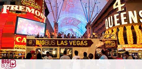 Image taken at dusk showing a bustling Fremont Street in Las Vegas with bright neon signage. A crowd is gathered, and in the foreground, a Big Bus tour with passengers on the upper deck can be seen. The dazzling lights of the overhead screen and surrounding casinos create a vivid atmosphere.