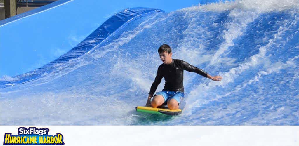 A person in a black shirt and blue shorts is surfing on a wave simulator. The artificial wave is pictured with a blue and white water pattern. In the background, the Six Flags Hurricane Harbor logo is visible, suggesting the location is a water park. The sky above appears clear and sunny.