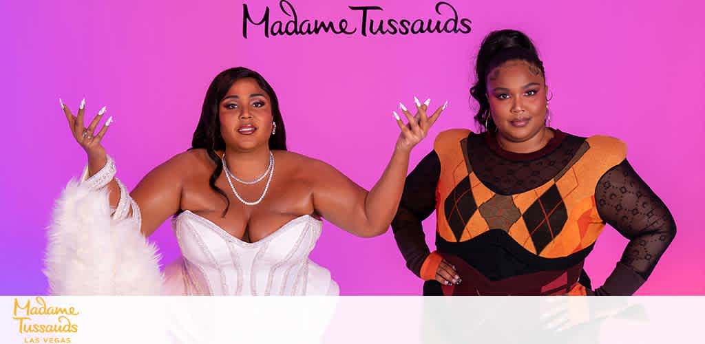 Image displaying two figures against a vibrant pink background with the Madame Tussauds Las Vegas logo. The figure on the left is dressed in a white gown adorned with feathers and sparkling jewelry, while striking a playful pose with one hand raised. The figure on the right is wearing a colorful geometric patterned outfit with sheer details, also posing elegantly with a hand lifted. Both figures exhibit intricate detailing and lifelike appearance.