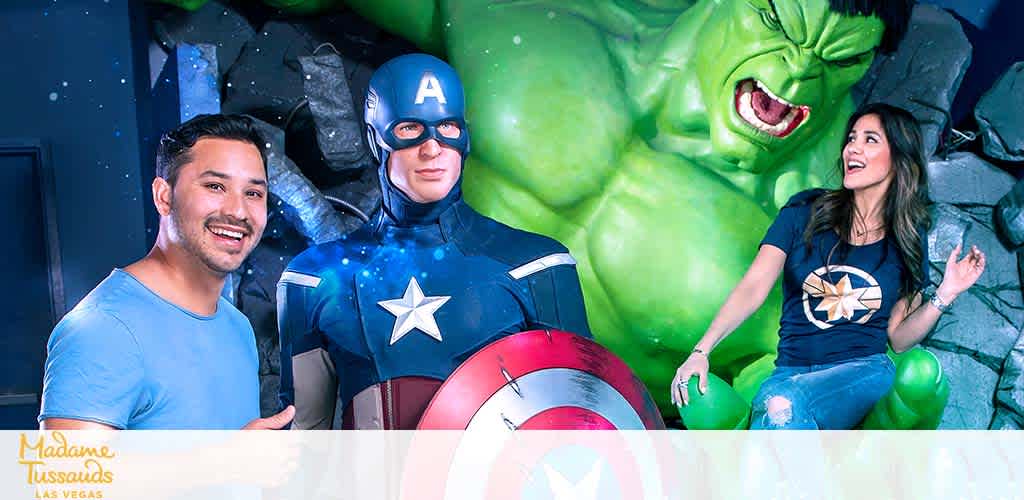 Image from Madame Tussauds Las Vegas showing a man and a woman posing with lifelike statues of Captain America and the Hulk. The man is to the left, smiling beside Captain America, who is holding his shield. The woman is seated to the right, looking thrilled next to the Hulk bursting through a wall. They're both wearing superhero-themed shirts.