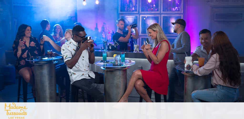 Image shows a vibrant social scene at Madame Tussauds in Las Vegas. Multiple individuals are engaging in various activities around high-top tables lit by blue hues. A man in a white shirt snaps a photo, while a woman in a red dress smiles at her phone. Drinks and laughter appear abundant in this lively bar setting.