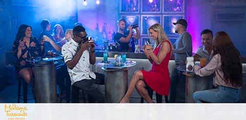 Image shows a lively bar scene with a diverse group of people. Some are chatting, while a woman in a red dress at the center looks away from a man who's holding a phone, possibly taking her photo. The ambiance is dimly lit with blue tones, giving a relaxed nighttime mood.