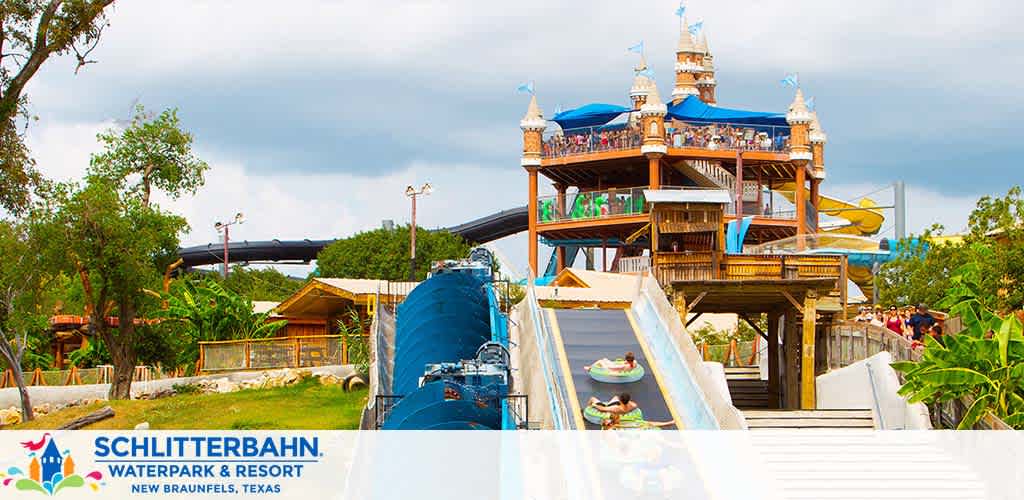 The image shows Schlitterbahn Waterpark and Resort in New Braunfels, Texas. Visitors are enjoying colorful water slides and attractions in an outdoor setting. A large water slide with a blue tube and an open-air slide with people on rafts descends from a tall, castle-like structure bustling with guests. Greenery surrounds the area under a partly cloudy sky.