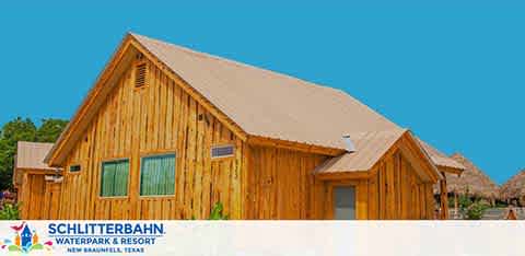 Image of a wooden cabin with a steep gabled roof under a clear blue sky. The cabin is part of Schlitterbahn Waterpark and Resort in New Braunfels, Texas, as indicated by the logo on the bottom left corner. The structure's natural wood finish stands out against the bright sky, suggesting a warm and inviting resort setting.