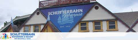 Image of Schlitterbahn Waterpark entrance featuring its blue and white signboard with a wave logo on a building that mimics German architectural elements. The establishment is surrounded by a cloudy sky overhead and advertises as a family-friendly destination in New Braunfels, Texas.