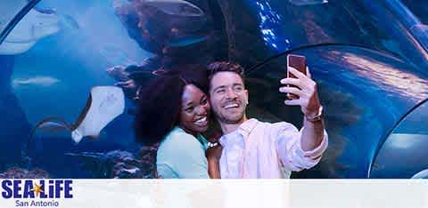 A joyful couple is taking a selfie at SEA LIFE San Antonio. They're in front of a large aquarium window with various fish in the background. Both are smiling warmly, with the woman embracing the man from behind. The SEA LIFE logo is visible in the lower-left corner, indicating the location.