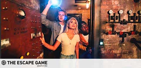 Three individuals are collaboratively exploring a room with rustic, industrial decor—evidently part of an escape game. The walls are adorned with dials and switches. One person holds a flashlight above, illuminating the scene as they search for clues, reflecting engagement and concentration. Their expressions convey a mix of intrigue and determination. The ambiance suggests adventure and teamwork as they navigate the challenge. The Escape Game logo is visible, indicating the experience's branding.