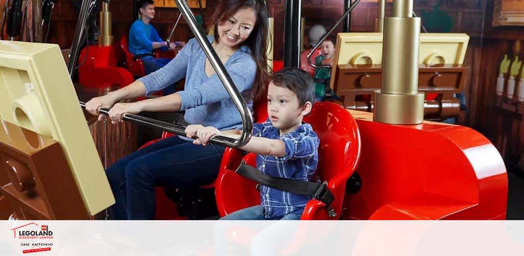 A joyful child and a smiling woman enjoy a ride in a bright red, LEGO-themed car at LEGOLAND San Antonio. They're harnessed in safely, ready for fun. Multiple rides and vibrant colors fill the background, capturing the excitement of the amusement park.