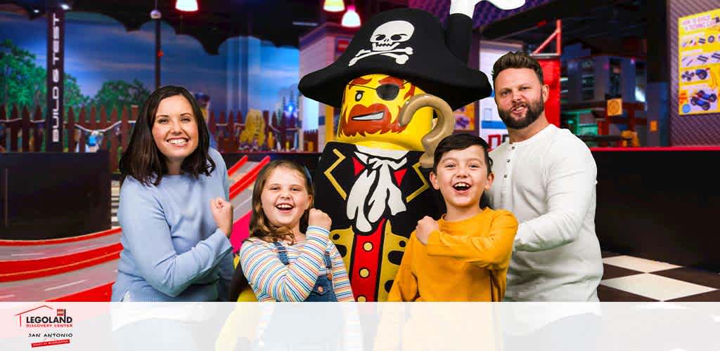 A family of four, with a smiling man, woman, girl, and boy, pose beside a life-sized LEGO pirate figure at LEGOLAND San Antonio. The pirate has a black hat, red coat, and a skull-and-crossbones design. They are all standing in front of a colorful amusement park backdrop with a racing track and LEGO scenery. The LEGOLAND logo is visible in the corner.