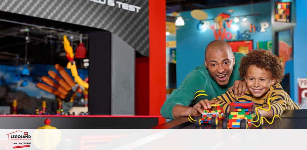 Image showing a smiling adult and child engaged in play at a Legoland Discovery Center in San Antonio. They are at a table with a colorful Lego creation in front of them. In the background, bright Lego structures and signage add to the playful setting. The logo of Legoland San Antonio is visible in the bottom left corner.