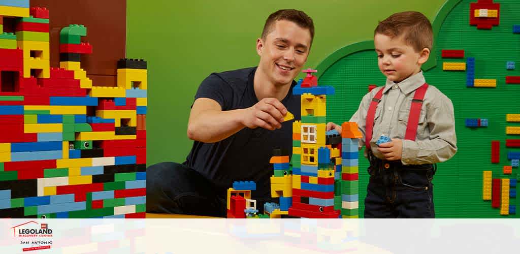 An adult and a child are engaging with LEGO blocks in a brightly colored room. Behind them, a wall adorned with multicolored LEGO pieces adds to the cheerful atmosphere. The adult, smiling, points at a LEGO creation while the child, in a shirt and suspenders, holds a LEGO piece, appearing curious and attentive. The logo of LEGOLAND San Antonio is visible, suggesting a playful and creative environment.