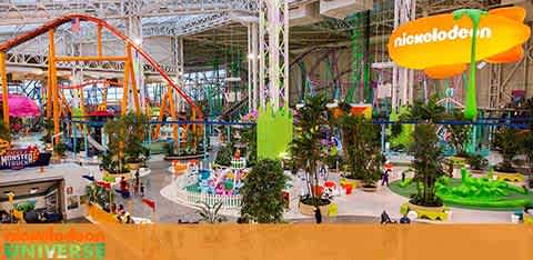 Image shows an indoor amusement park with vibrant orange and green roller coasters and various attractions. Central space features a carousel and a green seating area. Banners with Nickelodeon branding are visible. The atmosphere is lively with visitors enjoy the facility.