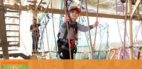 A child in a helmet and harness beams with joy on an indoor ropes course at Nickelodeon Universe. In the background, an adult participant and colorful structural elements are visible. The environment suggests adventure and fun.