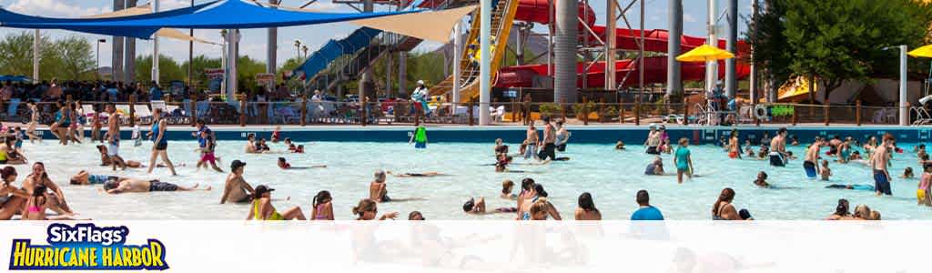 Visitors enjoy a sunny day at Six Flags Hurricane Harbor water park. Many are floating and swimming in a large wave pool. In the background, there are water slides, umbrellas, and lounging areas. The atmosphere is lively and summery.