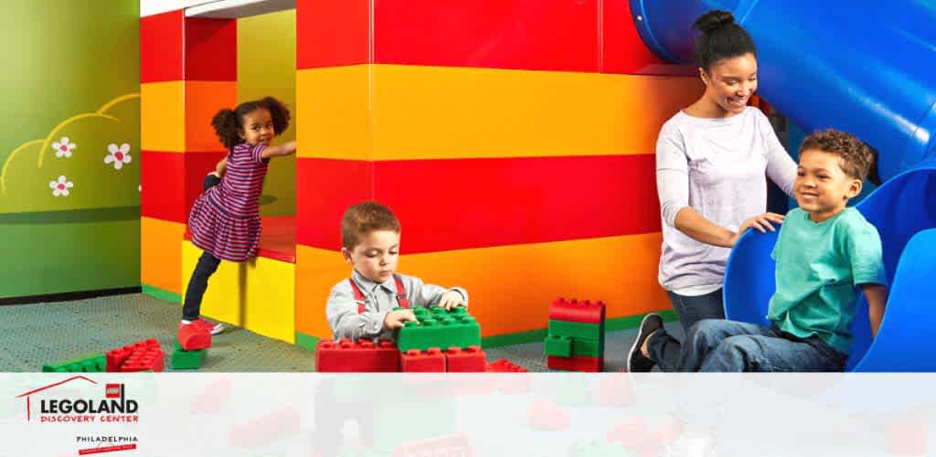 Image shows a bright and colorful play area in Legoland Discovery Center Philadelphia. A young girl in striped attire climbs a LEGO structure, a boy in red focuses on building with large LEGO bricks, and a smiling woman assists a cheerful boy seated in a blue area. Large LEGO pieces and vibrant decor enhance the playful atmosphere.