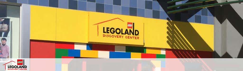 Image of Legoland Discovery Center facade with yellow signage and logo, adorned with large colorful Lego brick designs at the base. Bright sunlight casts shadows on the structure, and a person is partially visible on the left.