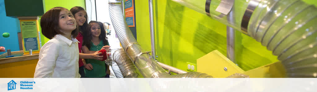 Image shows a group of children engaging with an exhibit at the Children's Museum Houston. They are watching with curiosity as an object travels through a clear, spiral tube. The environment is vibrant with green walls complementing the excitement on the children's faces.